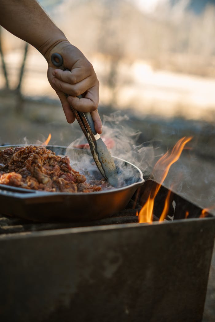 A hand preparing meat in a pan over an open fire, capturing the essence of rustic outdoor cooking.